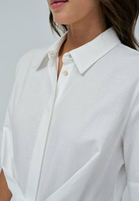Camicia bianca con colletto strutturato, realizzata in tessuto testurizzato. Presenta una pattina anteriore con bottoni chiari e maniche ampie.