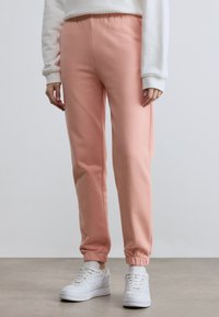 Calças de fato de treino cor-de-rosa com cintura e punhos elásticos, feitas de tecido macio. Combinadas com ténis brancos. O fundo é neutro e minimalista.