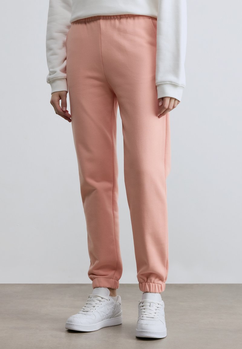 Calças de fato de treino cor-de-rosa com cintura e punhos elásticos, feitas de tecido macio. Combinadas com ténis brancos. O fundo é neutro e minimalista.