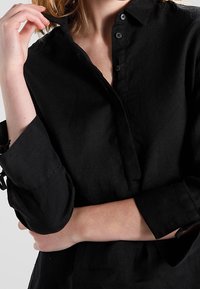 Sort linnedragt med knappet krave, lange ærmer og optrukne manchetter. Har en afslappet pasform og subtil struktur.