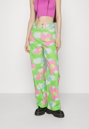 Persoon die groene broek draagt met grote roze bloemen, een roze cropped mouwloos topje en zwarte platformschoenen tegen een effen witte achtergrond.