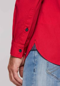 Rotes Langarmhemd mit strukturiertem Finish, ausgestattet mit dunklen Knöpfen an den Manschetten und einem abgerundeten Saum, kombiniert mit hellblauen Jeans.