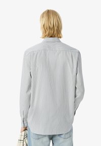 Una persona con capelli biondi di lunghezza spalla indossa una camicia a righe e tiene una sciarpa a quadri, vista da dietro in un contesto neutro.