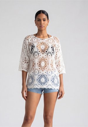 Vrouw met donker haar in een witte bloemrijke kanten top over een zwarte bikini en blauwe denim shorts, staand tegen een eenvoudige lichte achtergrond.
