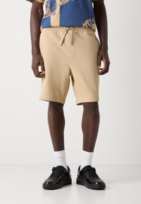 Shorts en coton beige avec taille élastique, poches latérales et texture lisse. Assortis avec des baskets noires et des chaussettes blanches montantes.