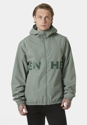 Mężczyzna z tatuażami ubrany w szałwiowozieloną bluzę z kapturem na zamek, z ciemnozielonym fragmentem tekstu, stojący na jednolitym jasnym tle.