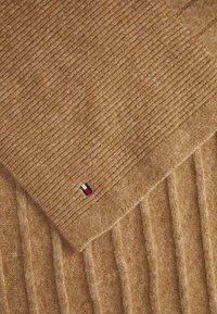 Beige knitted scarf featuring ribbed texture, soft wool material, and a small logo in red, white, and blue at the corner.