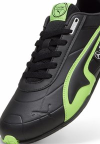 Sneaker de sport noir en matériau synthétique lisse, avec des accents verts, une semelle texturée et un détail de marque bien visible sur la languette et sur le côté.