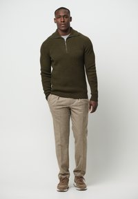 Olivgrüner gerippter Pullover mit einem halben Reißverschlusskragen, kombiniert mit beigen maßgeschneiderten Hosen und braunen Sneakers. Einfaches, strukturiertes Design über die gesamte Fläche.