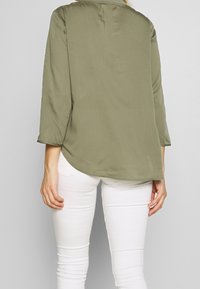 Blusa leggera verde oliva con maniche a tre quarti, orlo leggermente curvo e una lucentezza sottile, indossata con jeans bianchi aderenti.