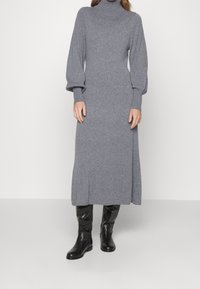 Šedé pletené šaty s dlouhým rukávem, vysokým límcem, volným střihem a rovnou siluetou, doplněné černými koženými vysokými botami po kolena.