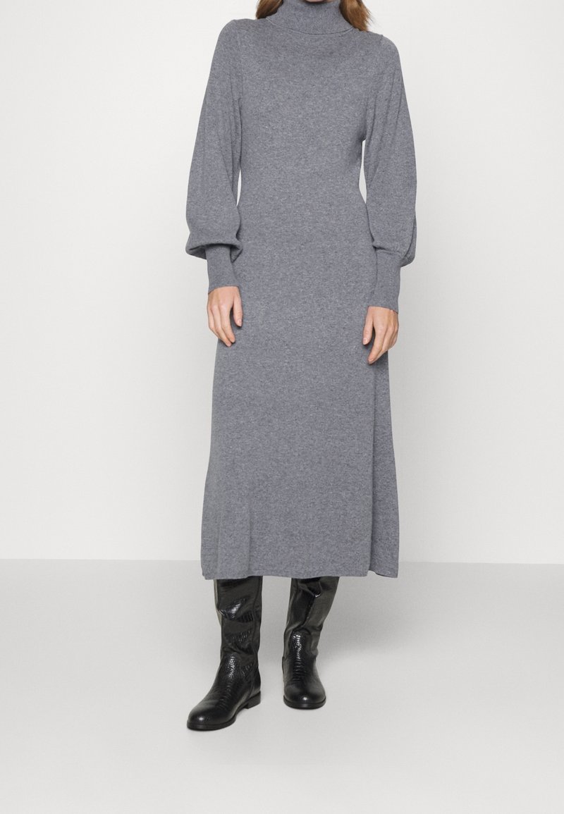 Šedé pletené šaty s dlouhým rukávem, vysokým límcem, volným střihem a rovnou siluetou, doplněné černými koženými vysokými botami po kolena.