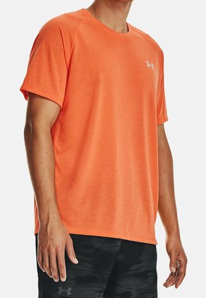 Mann trägt ein leuchtend orangefarbenes, kurzärmeliges Sport-T-Shirt und schwarze gemusterte Shorts mit Logo auf der Brust und am linken Oberschenkel.