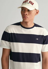 Gestreepte t-shirt in marineblauw en roomkleurig katoen met een ronde hals. Bevat een klein geborduurd logo op de borst. Gemaakt in combinatie met een beige pet.