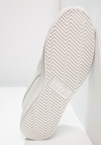 Vit sneakersula med ett fiskbensmönster, texturerad kant och en logotyp präglad på sidan. Slät yta med subtila detaljer.