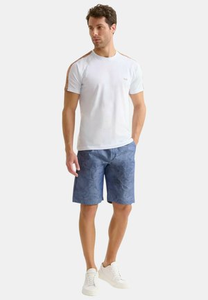 Outfit casual da uomo con una maglietta azzurro chiaro con dettagli marroni e pantaloni corti blu a fantasia, abbinati a sneakers bianche.