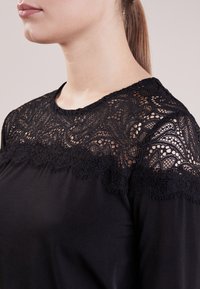 Schwarzes Oberteil mit einem Spitzenüberwurf an den Schultern im wellenförmigen Muster. Glatter Stoffkörper mit Rundhalsausschnitt und kurzen Ärmeln.