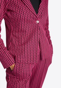 Roze-zwart patroonblazer met een aangesloten pasvorm, enkele knoopsluiting en twee zijzakken; het materiaal lijkt textuur en een geweven structuur te hebben.