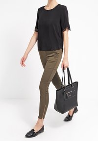 Svart kortärmad topp med spetsdetaljer, olivgrön figurnära byxor med dragkedjedetaljer, svart axelväska och svarta loafers.