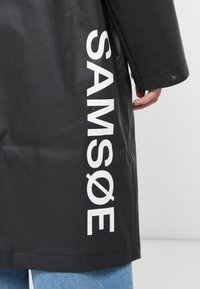 Black waterproof coat with a shiny finish; features large white text reading "SAM SøE" along the left side; straight cut and button details.