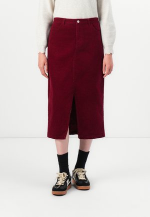 Mid-lange bordeauxrode rok met een split aan de voorkant, gedragen met zwarte sokken, zwart-witte sneakers en een lichtbeige top met lange mouwen.