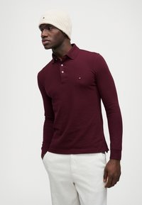 Pólo de manga longa de cor bordô com placket de três botões e pequeno detalhe de logotipo. Usado com calças de cor clara e um gorro de malha creme.