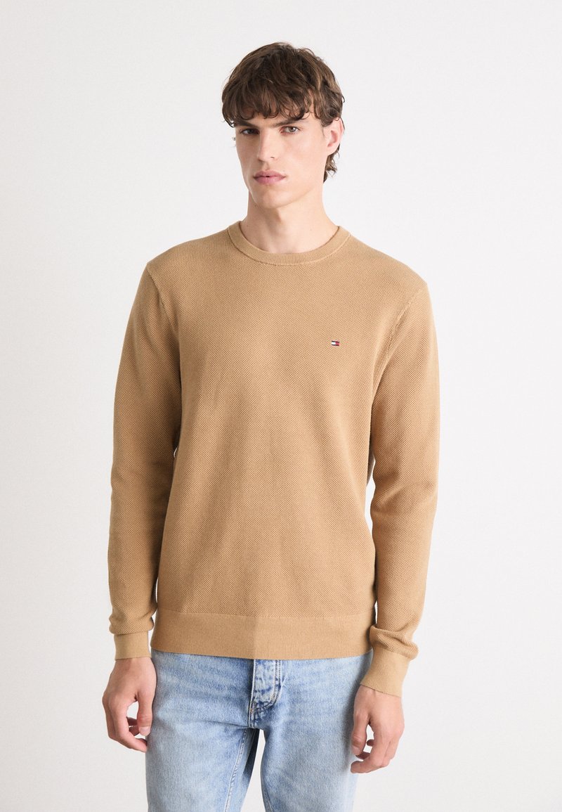 Maglione beige a maglia con maniche lunghe e collo rotondo. Presenta una texture sottile e un piccolo logo sul petto. Abbinato a jeans azzurri chiari.