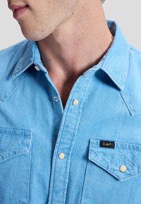 Homme portant une chemise en denim bleu clair avec des boutons-pression en nacre et une étiquette de marque Lee sur la poche poitrine, cou et bas du visage visibles.