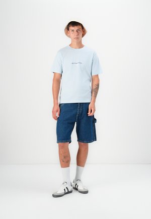 Világoskék, rövid ujjú póló, amelyen a "JACK & JONES" logó található, kék farmer rövidnadrággal és szürke sportcipővel párosítva, fehér és fekete részletekkel.