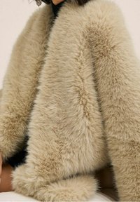 Femme portant un manteau en fausse fourrure beige doux sur une tenue noire, montrant la texture et un style confortable des épaules jusqu'au milieu des cuisses.