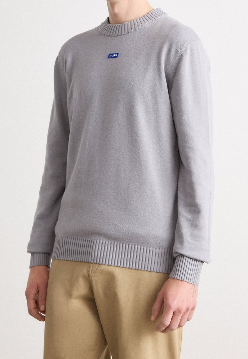 Homme portant un pull gris clair à manches longues avec col, poignets et ourlet côtelés, associé à un pantalon beige, debout devant un fond blanc.
