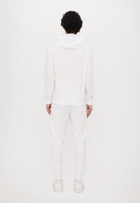 Hvid hættetrøje og matchende sweatpants, der har lange ærmer, snoretræk på hætten, manchetkanter og flade sneakers. Glat bomuldsstruktur.