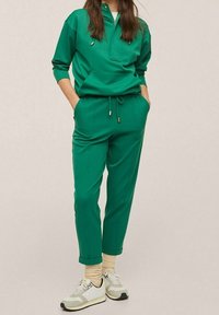 Groene sportieve set met een top met rits en joggingsbroek met touwtjes, gemaakt van gladde stof. Gecombineerd met lichtgekleurde sneakers.