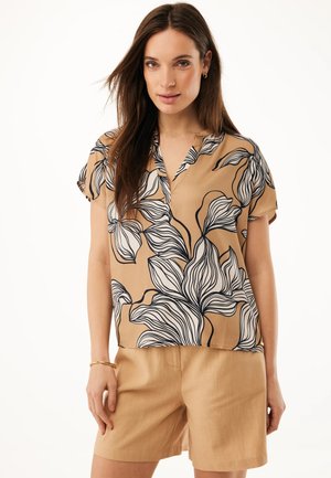 Kvinde iført beige shorts og en beige kortærmet top med fedt sort og hvidt blomsterprint, stående mod en hvid baggrund.