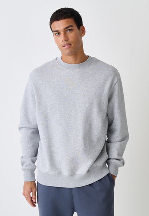 Jonge man met kort donker haar, die een lichtgrijze trui met ronde hals en donkere broek draagt, staat voor een effen witte achtergrond.