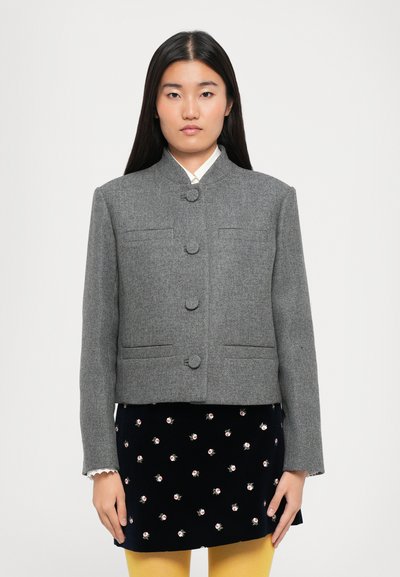 Veste grise courte en tissu texturé, avec un col montant, quatre boutons sur le devant et deux poches latérales. Portée par-dessus une jupe à fleurs.