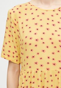 Κίτρινο φόρεμα με κοντά μανίκια, διακοσμημένο με ένα σχέδιο από μικρά ροζ λουλούδια με σκούρα κέντρα και ζώνη στη μέση.