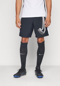 Čierne atletické šortky s bielym logom Nike, v kombinácii s čiernymi ponožkami po kolená a čiernymi kopačkami s modrými akcentmi. Hladká textúra látky.