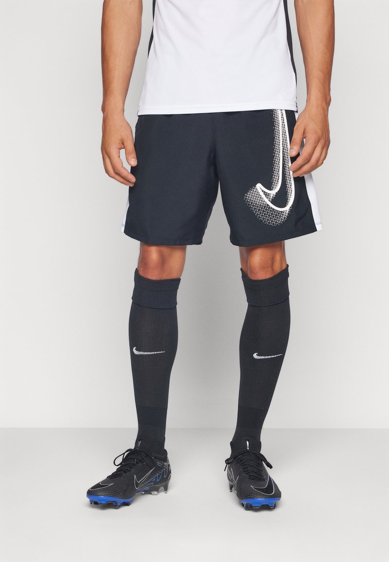 Čierne atletické šortky s bielym logom Nike, v kombinácii s čiernymi ponožkami po kolená a čiernymi kopačkami s modrými akcentmi. Hladká textúra látky.