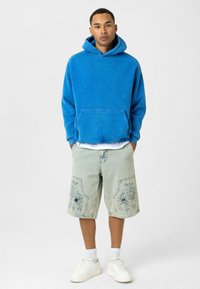 Blauwe hoodie met kangoeroezak, gecombineerd met lichte denimshorts met bloemenborduursels en versleten details, en witte sneakers.