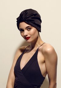 Svart sateng turbanlignende hodeplagg med en vridd knute foran. Glatt tekstur, tettsittende design, kombinert med en mørk, ermeløs antrekk.