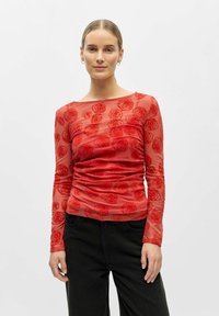 Sarkans caurspīdīgs tops ar garām piedurknēm, uz kura attēlots rozes zieds, ar savilktu tekstūru viduklī, kopā ar tumšām biksēm.