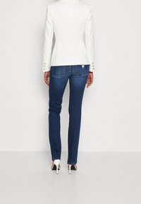 Blazer blanco ajustado con acentos de botones dorados, combinado con jeans ajustados azul oscuro y zapatos de tacón blanco, visto desde atrás.