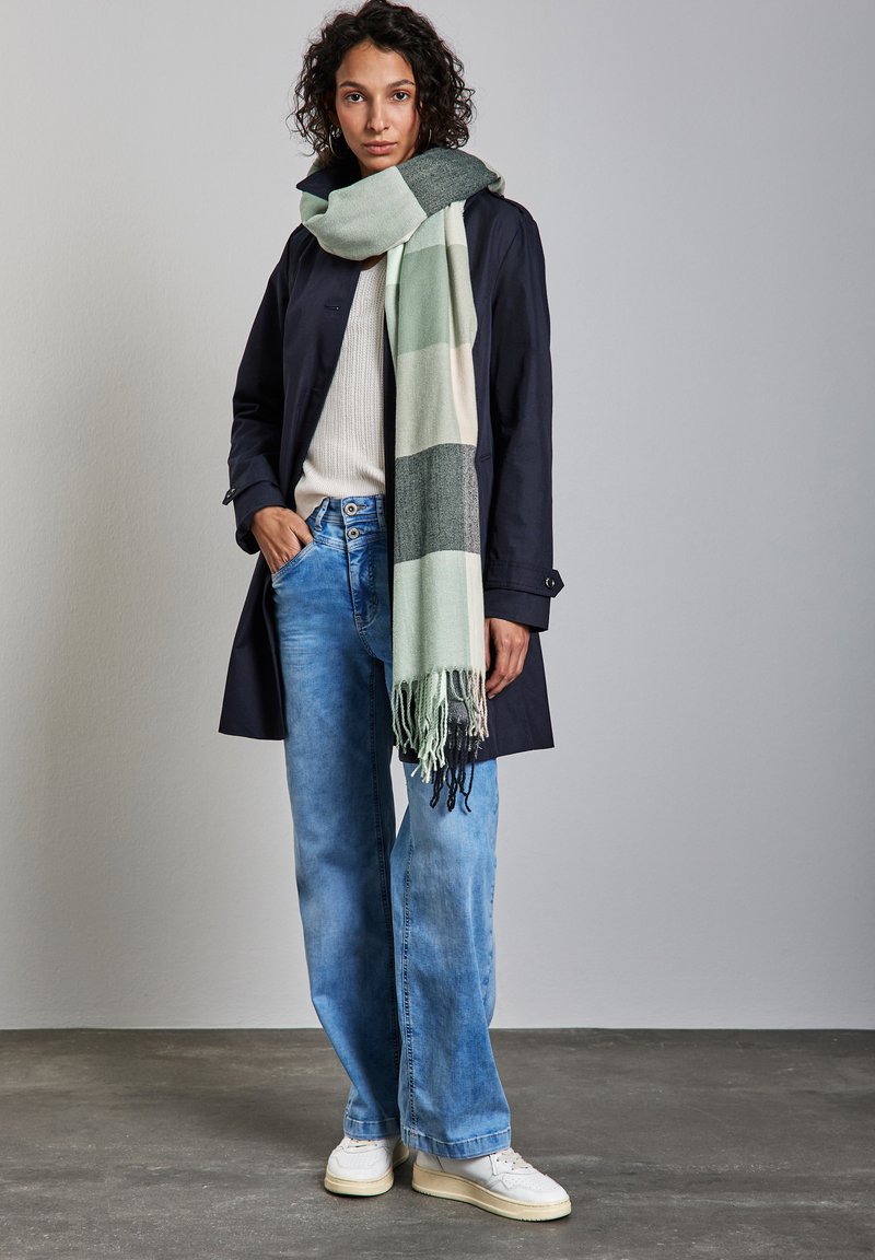 Manteau long en marine, pull blanc, jeans larges bleu clair, et une grande écharpe à carreaux verts et gris avec des franges, associée à des chaussures blanches.