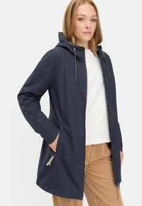 Veste à capuche bleu marine en tissu lisse avec une fermeture éclair complète, des poches latérales et des cordons de serrage réglables. Portée par-dessus un haut blanc et un pantalon beige.