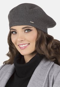 Szürke kötött beret, laza dizájnnal, fémbetét jellemzi. Fekete garbóval és szürke kabáttal viselve. Sima textúra.