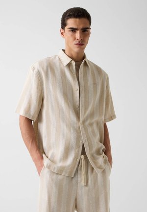 Homme portant une chemise à manches courtes boutonnée à rayures verticales beige et blanches, et un short assorti avec cordon de serrage, mains dans les poches, fond neutre.