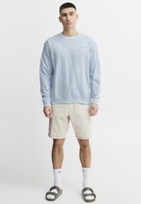 Felpa azzurra con collo a giro, polsini e orlo a costine. Abbinata a pantaloni corti beige e sandali verdi. Presenta un piccolo logo blu.