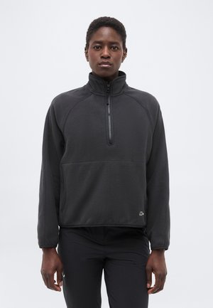 Άτομο που φοράει γκρι σκούρο φλις μπλουζάκι με φερμουάρ μέχρι τη μέση και μαύρο παντελόνι, στέκεται μπροστά σε ένα απλό ανοιχτό φόντο.