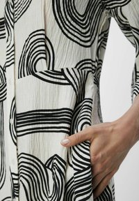 Robe noire et blanche à motifs présentant des lignes ondulées sur un tissu blanc texturé, avec une poche et des boutons près du col.